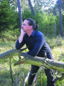  Photo von Rainer Wüst. Ein dunkelhaariger Mann um die 40 steht mit einer auf einem Zaun aufgestützten Hand im Wald und schaut nachdenklich in die Ferne.